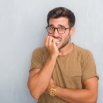 Handsome young man over grey grunge wall wearing glasses looking stressed and nervous with hands on mouth biting nails. Anxiety problem.