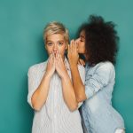 Young woman telling her girlfriend some secret. Two women gossiping. Excited emotional girl whispering to her friend ear, turquoise studio background