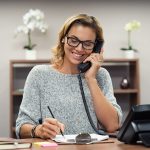 Beautiful mature woman talking on phone at creative office. Happy smiling businesswoman answering telephone at office desk. Casual business woman sitting at desk making telephone call and taking note.