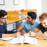 Three students studying and doing homework together. Young men talking, writing and sitting at desk in classroom or library. Education concept.