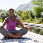 Portrait of smiling woman in lotus position near swimming pool looking at camera. Healthy black mature woman meditating and sitting in lotus pose on yoga mat. Happy lady in zen pose doing meditation.