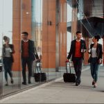 Business man and woman walking outside, holding coffee and rolling bags, talking