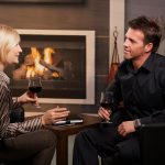 A man and woman discuss business in a hotel lobby over drinks, fireplace in the background