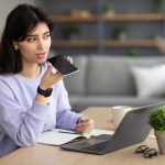 Woman sitting at a computer uses voice search assistant on smart phone