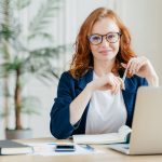 Female with red hair and glasses holds a pen while sitting at desk with laptop and notebook, smiling confidently and looking at the camera