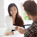 Confident Asian woman shakes male colleague's hand over laptop