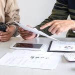 Close up of two men holding documents and coffee, with computers and smartphones nearby