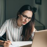 Young long-haired brunette woman in a horizontal striped shirt stares intently at laptop computer, pen in hand poised over notebook.