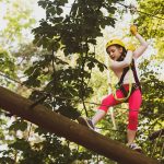 Child with yellow helmet, climbing gear, and harness steps out onto a log, high in the air