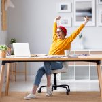 Woman sitting at a desk in front of her computer raises both arms with a smile on her face, after she let go of a client relationship that wasn't working out