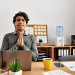 Young hispanic man wearing business style sitting on desk at office begging and praying with hands together with hope expression on face