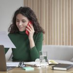 Young smiling spanish businesswoman reading paper document and talking via mobile phone