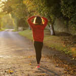 Woman in red sweatshirt, black leggins, and red sneakers walks in the early morning light on a long country road