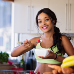 Athletic woman in exercise clothes smiles with a bowl of yogurt and fruit in a sunlit kitchen surrounded by bowls of fruits and vegetables