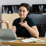 Professional woman gestures to her laptop in a persuasive way, notebook and glasses on her desk