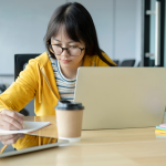 Woman in comfortable clothes and glasses makes notes off her laptop screen about conference attendees and information