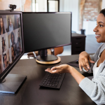 Professional woman attends virtual meeting in home office, gesturing to one of her two monitors, screen filled with other attendees