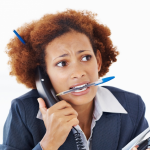 Harried woman holds telephone to her ear with arms full of papers and pen between her teeth, looking stressed
