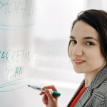 business woman stands at a whiteboard, marker in hand, drafting a client outreach roadmap