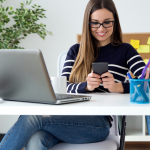 smiling writer checks her phone while sitting at her desk, laptop open and coffee mug nearby