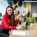 Smiling professional woman sits with co-workers at long table with computer and plants