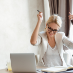 a woman raises her hands in triumph at her laptop