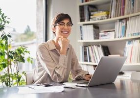 Successful young business woman sitting in creative office and looking at camera. Portrait of happy entrepreneur working on computer. Smiling businesswoman using laptop while working from home.