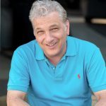 A seated, middle aged man of greek heritage with a warm, smiling face, wavy short grey hair, and  blue polo shirt.